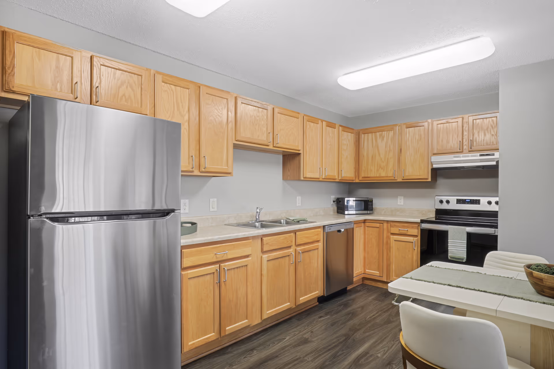 A modern kitchen with light wood cabinets, a stainless steel refrigerator, dishwasher, and electric stove. There is a double sink under a row of upper cabinets, a microwave on the countertop, and a small dining table with chairs in the foreground.