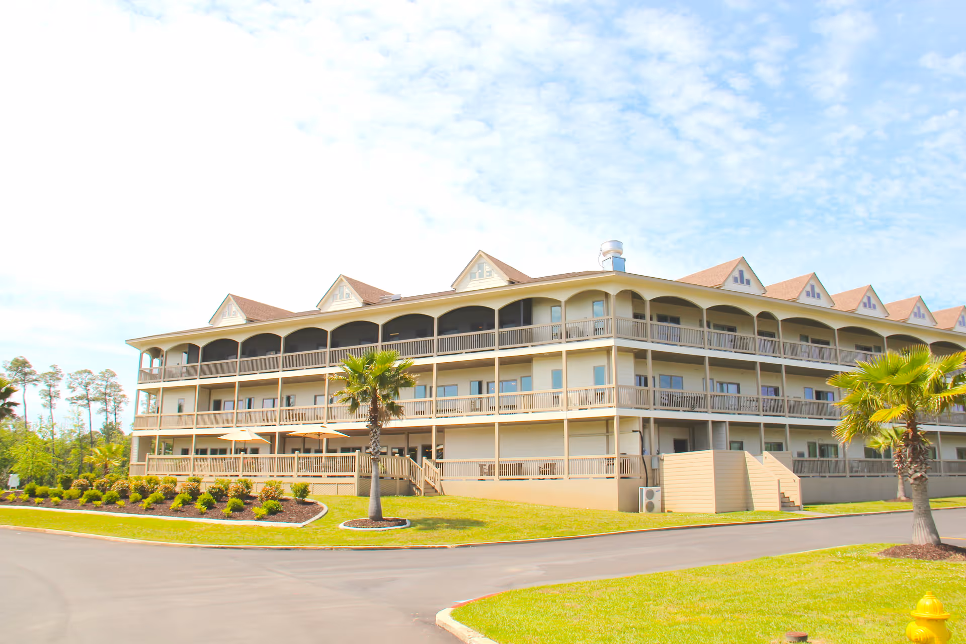 Exterior view of a large, three-story residential building with multiple balconies and a sloped roof, surrounded by landscaped greenery and palm trees under a partly cloudy sky.