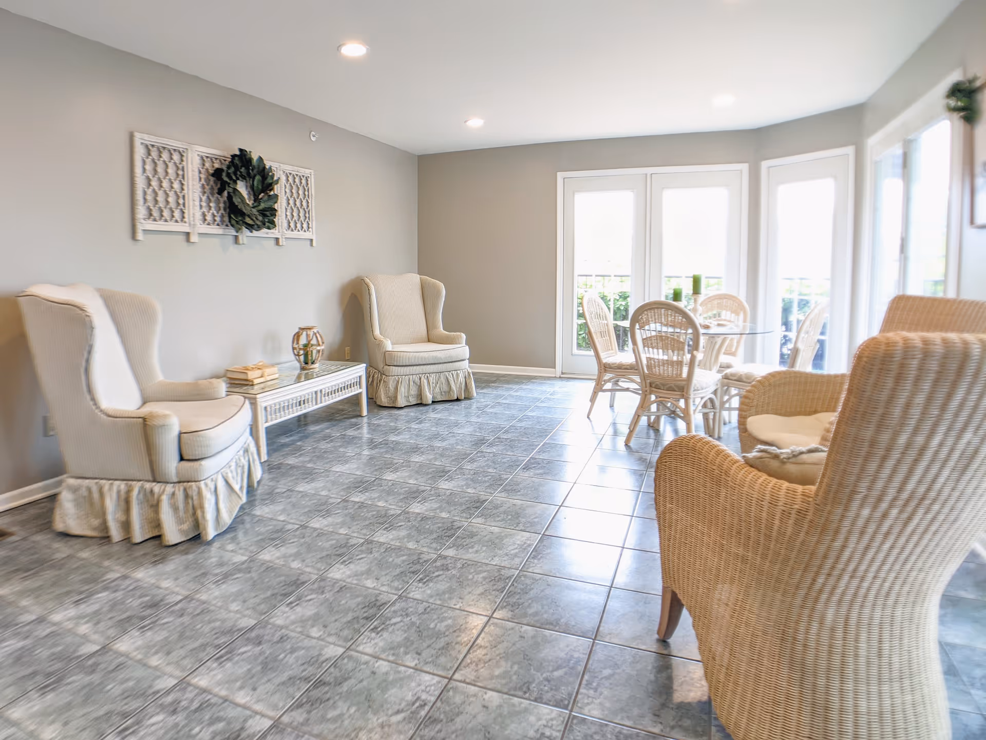 A bright and airy living room area with gray tiled flooring, two beige upholstered armchairs with skirts, a white coffee table with decorative items, and a round glass dining table surrounded by wicker chairs near large windows and glass doors.
