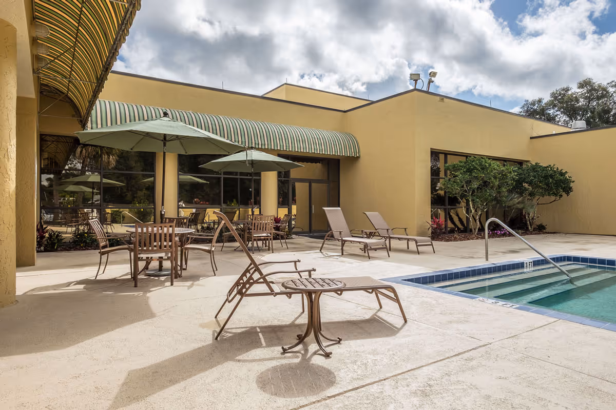 Outdoor patio area at Grand Villa of DeLand featuring several lounge chairs and tables with umbrellas next to a small swimming pool. The building has yellow walls and large windows with green and white striped awnings. The sky is partly cloudy.