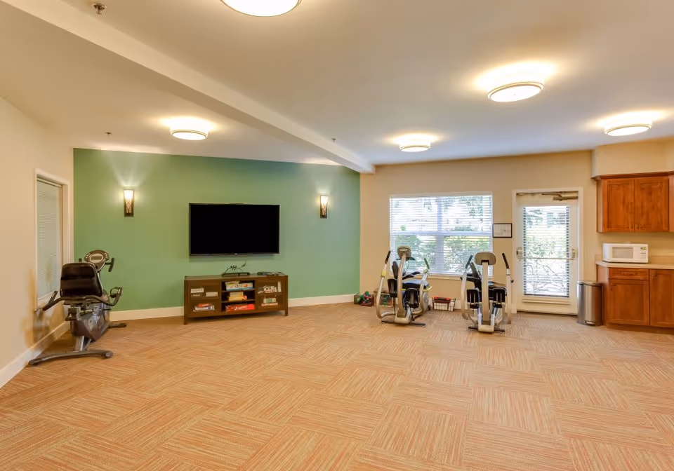 A spacious room with exercise equipment including stationary bikes and elliptical machines, a wall-mounted flat screen TV on a green accent wall, a wooden cabinet with board games, large windows letting in natural light, and a kitchenette area with wooden cabinets and a microwave.