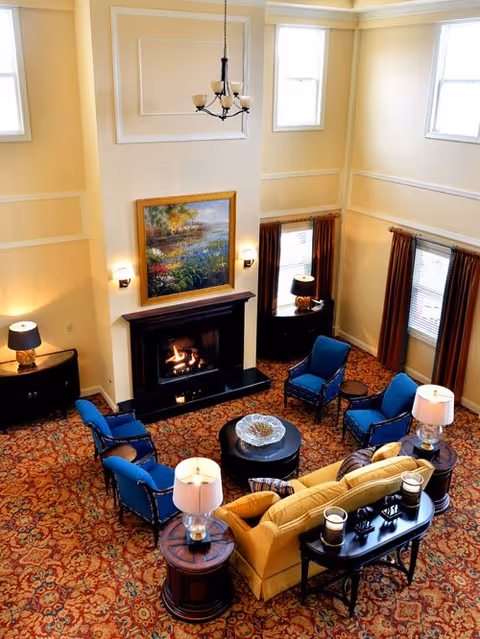 Two-story common living room with a fireplace, framed painting, a yellow sofa and blue armchairs arranged around a central coffee table on a patterned carpet.