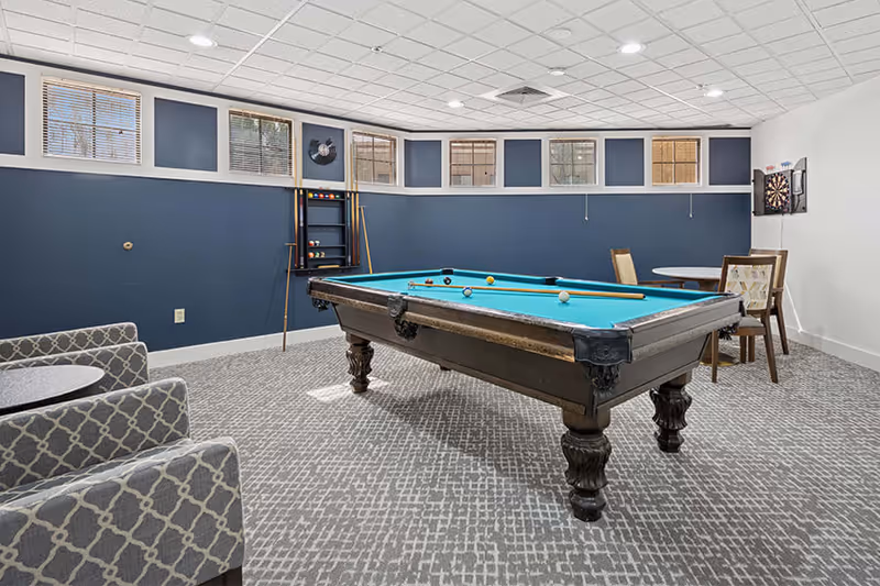 Carpeted recreation room with a pool table in the center, seating, a dartboard and small tables against navy blue walls.