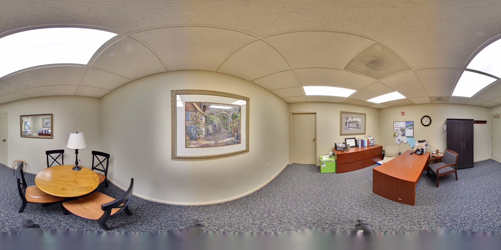 A senior living facility office area with a round wooden table and four chairs on the left side, a lamp on the table, a framed painting on the wall, and a desk with office supplies, chairs, a clock, and a cabinet on the right side. The room has a carpeted floor and a drop ceiling with fluorescent lights.