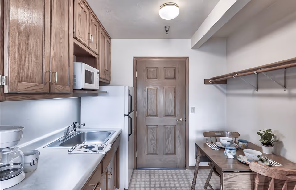 A small kitchen area with wooden cabinets, a white refrigerator, microwave, and sink. There is a coffee maker and a coffee mug on the counter. To the right, there is a wooden table set with bowls, cups, and utensils, along with two wooden chairs. A wooden door is centered in the background, and a shelf with hanging rods is mounted on the right wall.