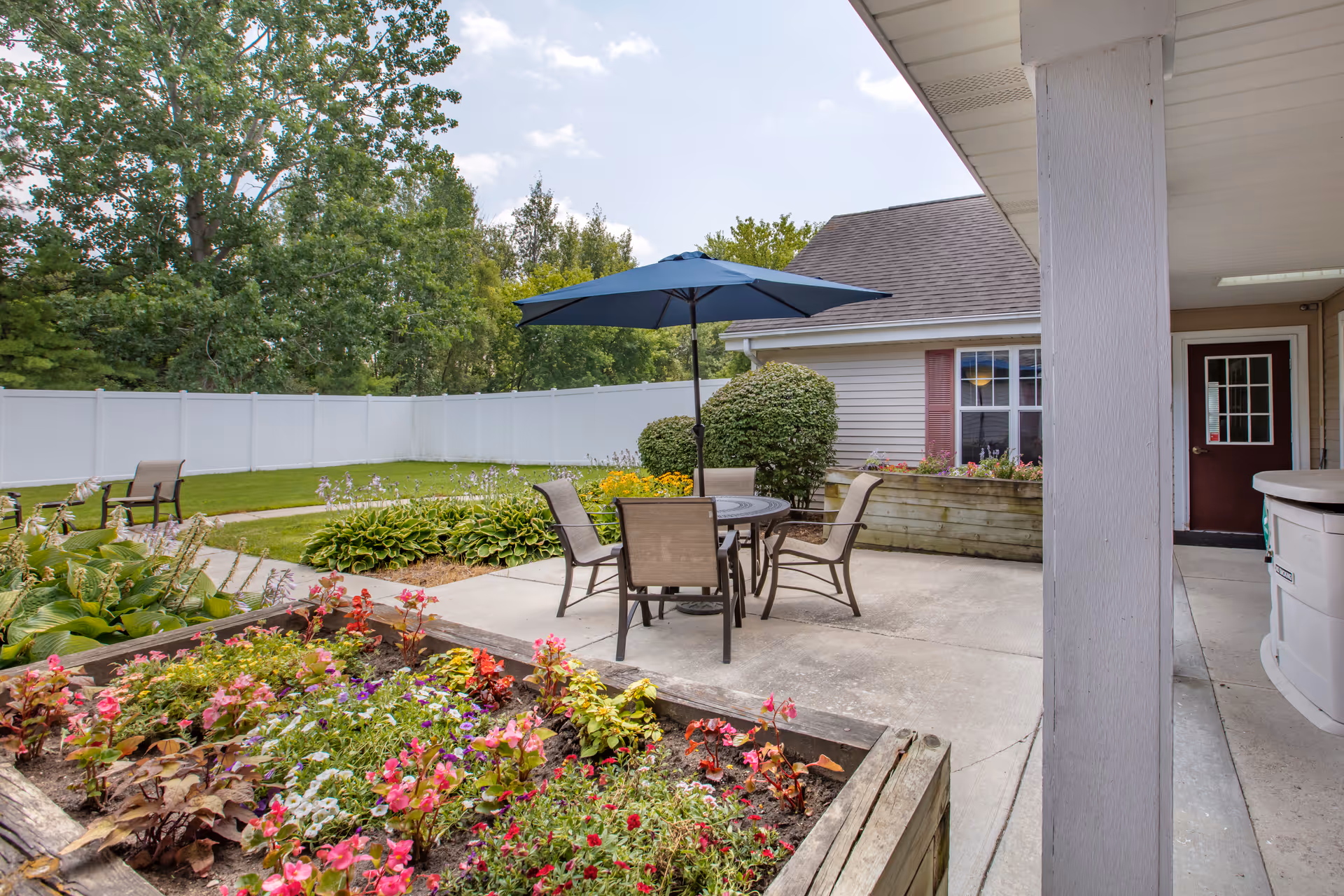 Outdoor patio area with a round table and four chairs under a blue umbrella, surrounded by flower beds and greenery. There is a white fence enclosing the area and a building with beige siding and a red door in the background.