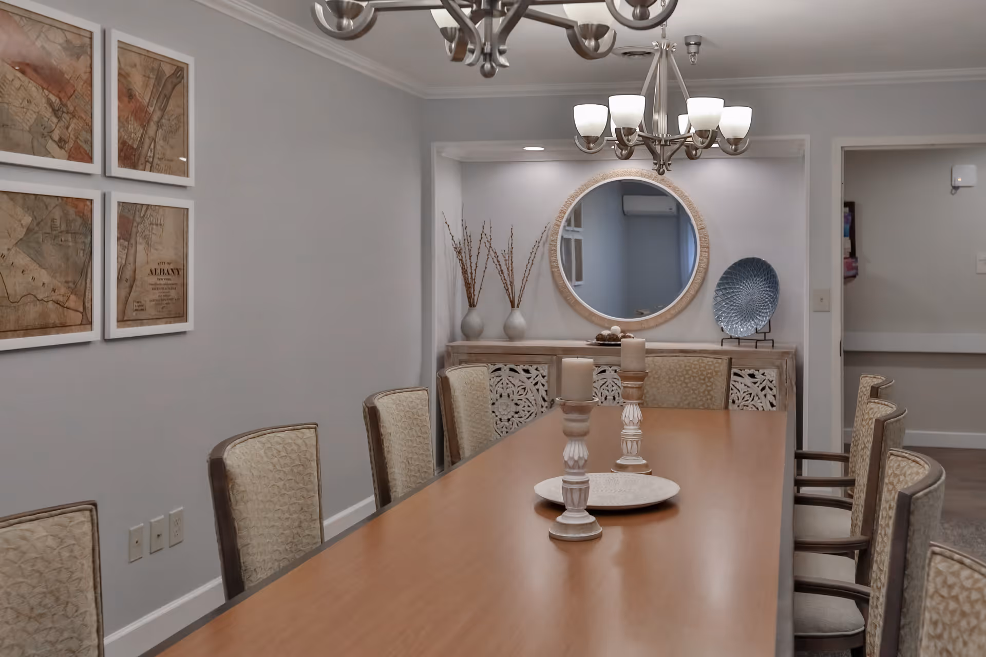 A dining room with a long wooden table surrounded by upholstered chairs. On the table are two decorative candle holders and a round tray. The wall features four framed vintage maps, and a sideboard with decorative vases and a round mirror above it. A chandelier with multiple lights hangs from the ceiling.