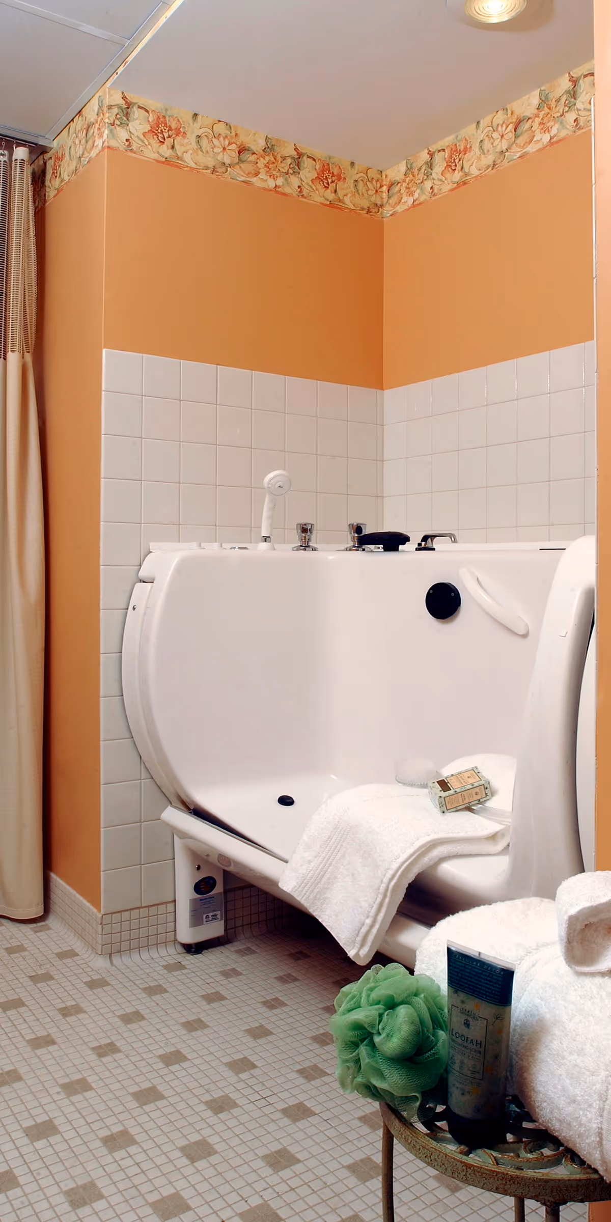 A bathroom corner featuring a walk-in bathtub with a white towel and soap placed on its seat. The walls are painted peach with a floral wallpaper border near the ceiling and white square tiles halfway up the wall. The floor has small square tiles in a beige and brown pattern. A small table nearby holds a green bath sponge, a tube of lotion, and additional white towels.