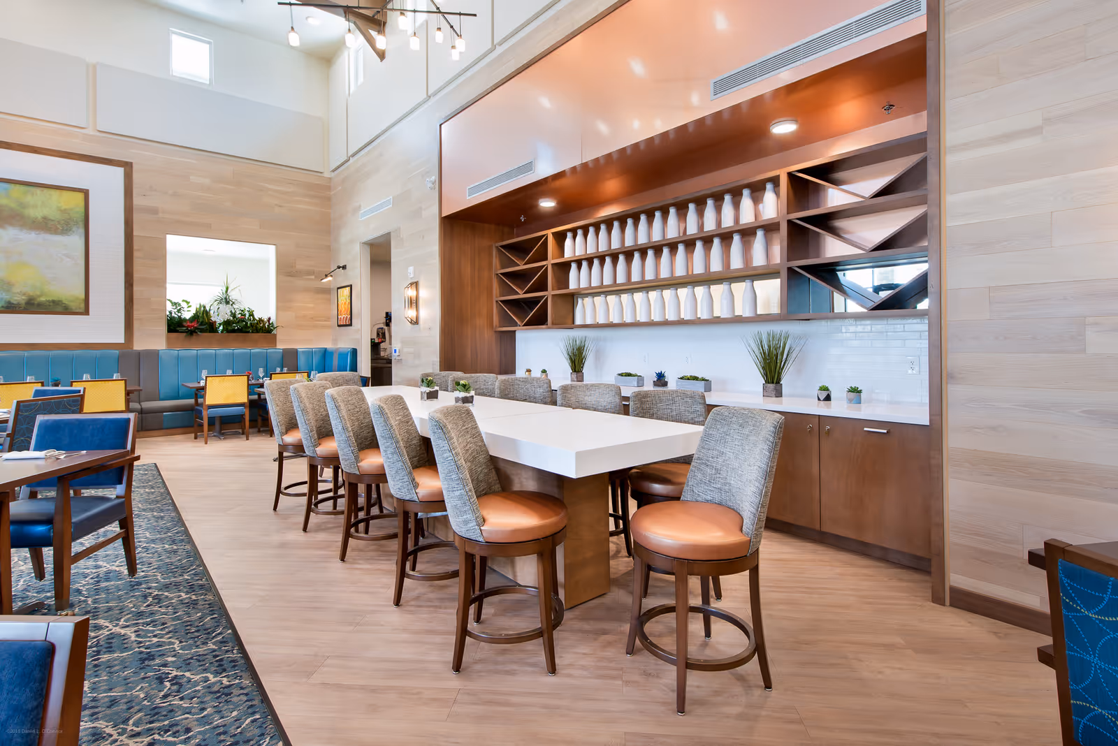A bright and modern dining room with a long white table surrounded by cushioned chairs with wooden legs. The back wall features wooden shelving filled with white decorative bottles and small potted plants on the countertop. Additional seating with blue and yellow chairs and a blue cushioned bench is visible along the left side. The room has light wood flooring, high ceilings with windows, and contemporary lighting fixtures.