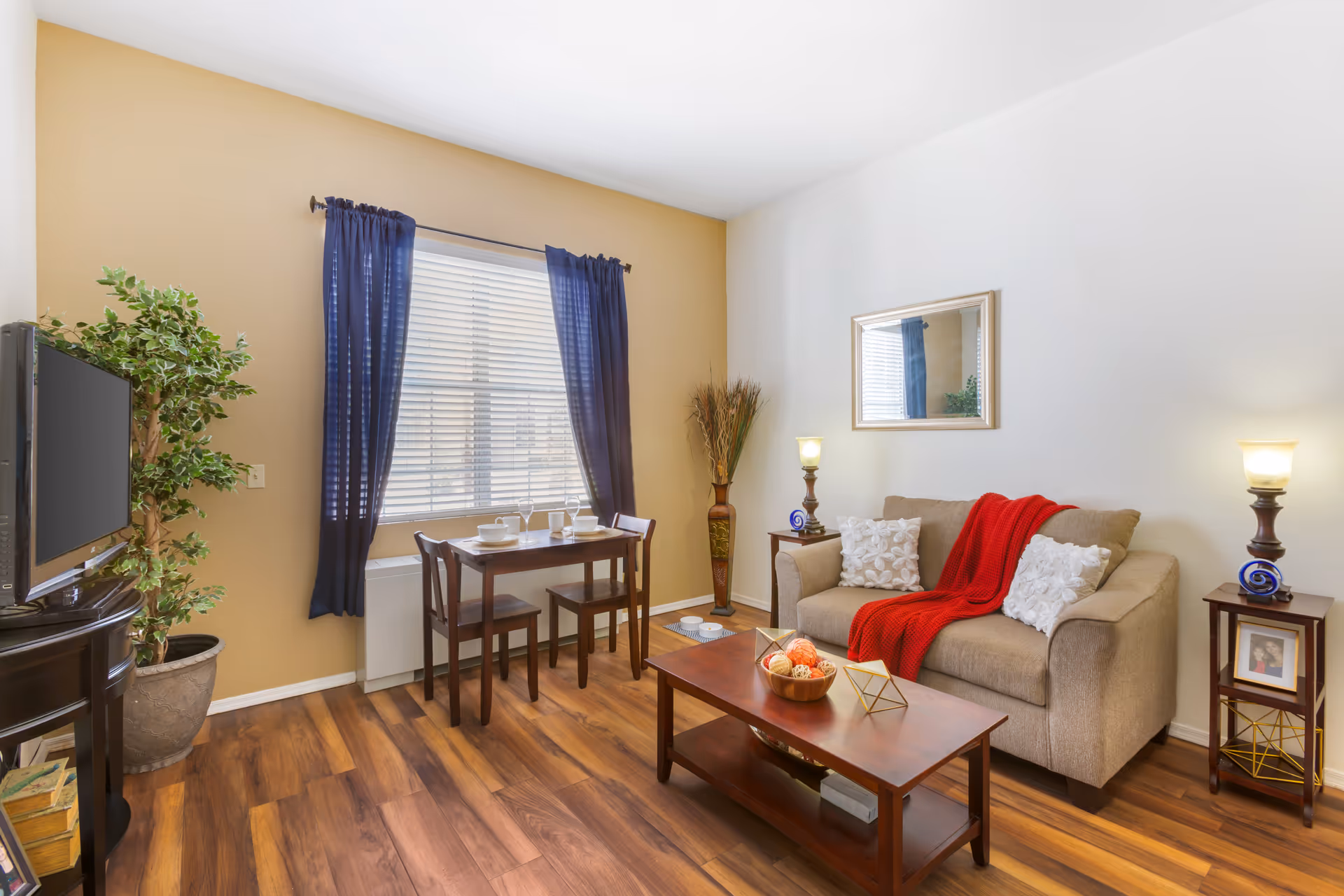 A cozy living room with a beige sofa adorned with white pillows and a red throw blanket. A wooden coffee table with decorative items sits in front of the sofa. There are two small side tables with lamps on either side of the sofa. A wooden dining table with two chairs is placed near a window with blue curtains. A flat-screen TV is positioned on a dark wooden stand next to a potted plant. The room has wooden flooring and light-colored walls, one painted beige and the other white.