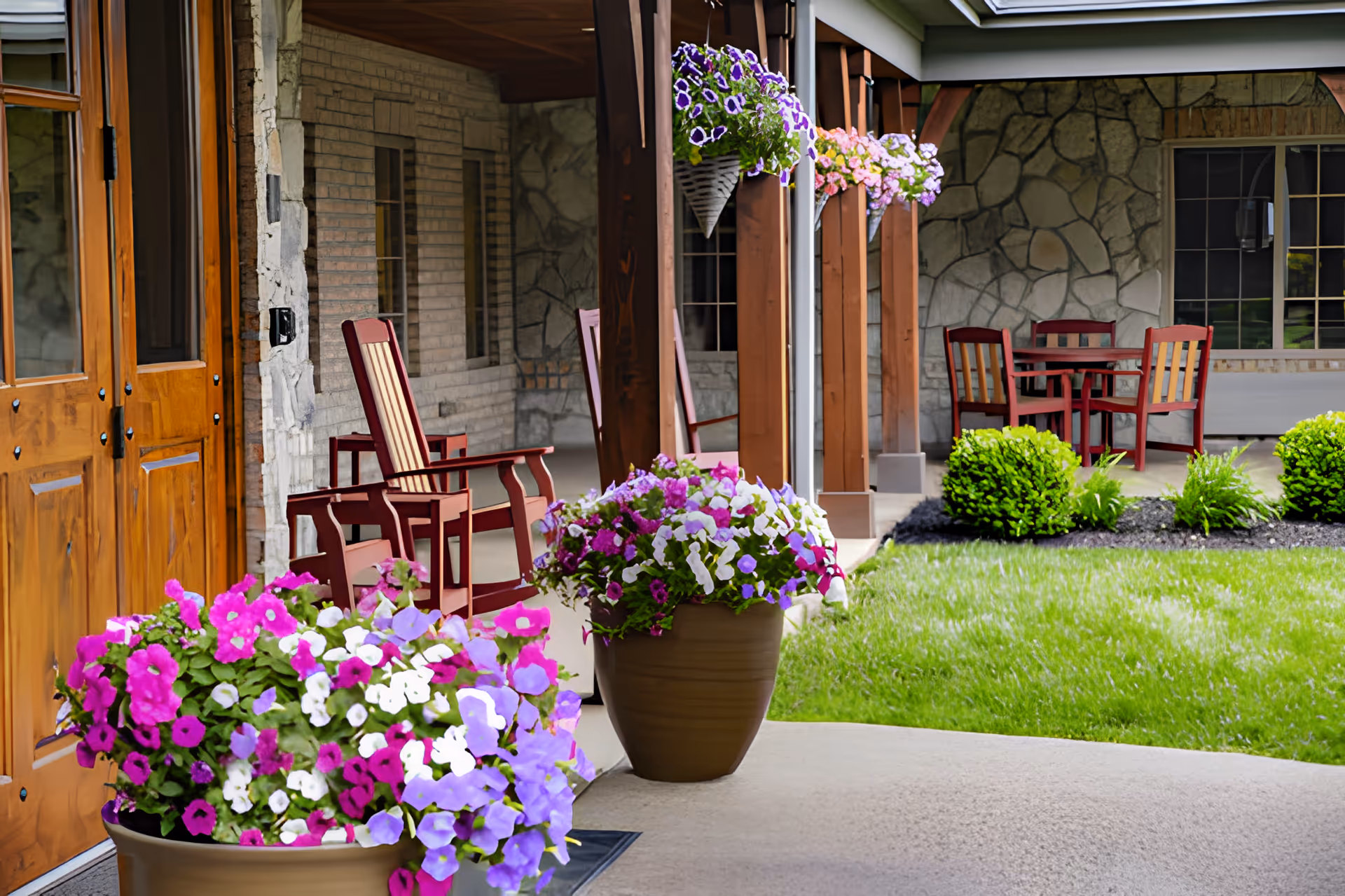 A covered outdoor patio area at The Inn at Walnut Trail featuring wooden rocking chairs, a table with chairs, hanging flower baskets, and large flower pots with vibrant pink, purple, and white flowers. The patio is adjacent to a stone and brick building with wooden doors and windows, overlooking a well-maintained grassy lawn with shrubs.