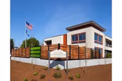 Exterior view of Columbia Lutheran Home building with a modern design, featuring large windows and a wooden fence. An American flag is flying on a flagpole to the left of the building under a clear blue sky.