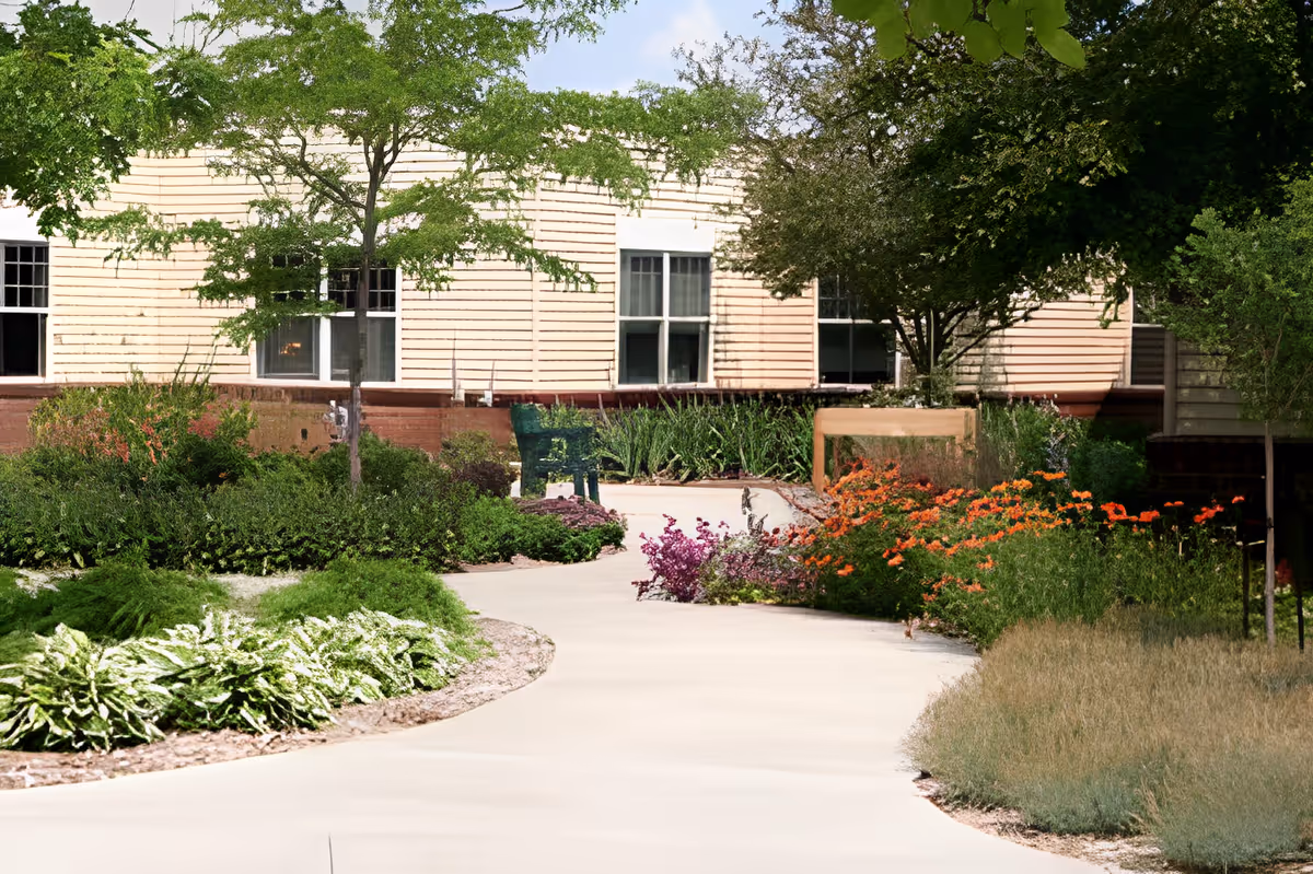 A winding concrete pathway surrounded by lush greenery and colorful flowers leading to a beige building with multiple windows. Trees and shrubs line the path, creating a serene garden-like outdoor space.