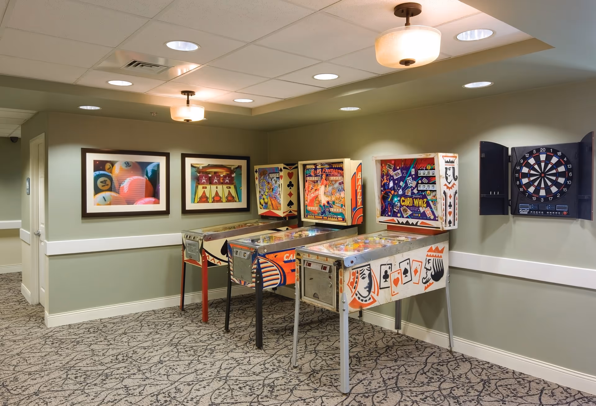 A small indoor game room with three vintage pinball machines, framed pictures on the wall, a closed dartboard cabinet, and ceiling lights.