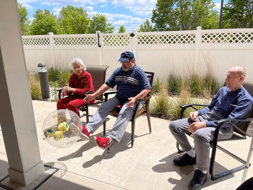 Three elderly individuals sitting outside on patio chairs in a fenced area with greenery behind them. One man in the center is kicking a large transparent ball with yellow blocks inside. The weather is sunny with a blue sky and some clouds.