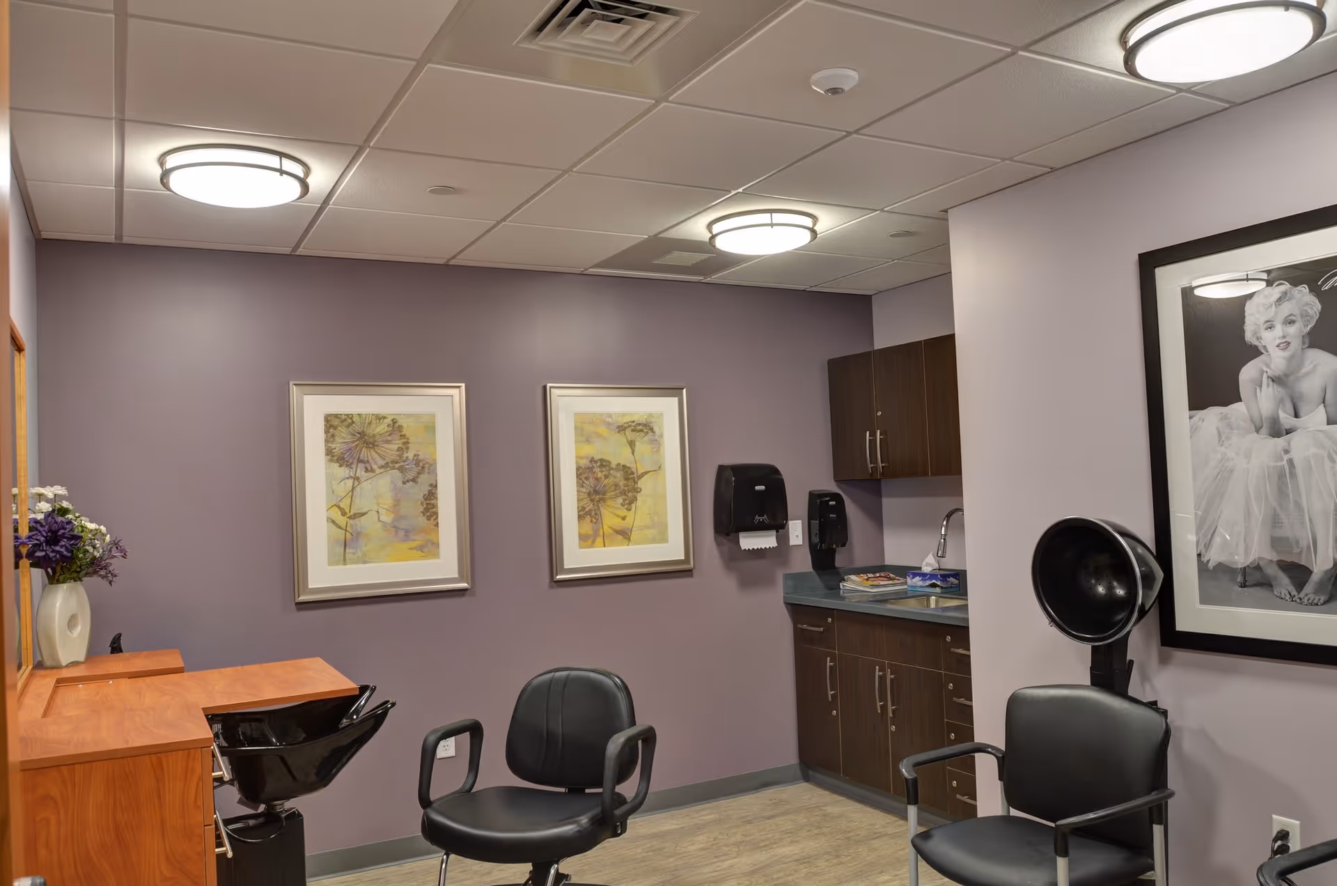Interior of a salon room with two black salon chairs, a hair washing station, wooden cabinets, a sink, two framed floral artworks on a purple wall, a black and white framed photo of a woman, and ceiling lights.