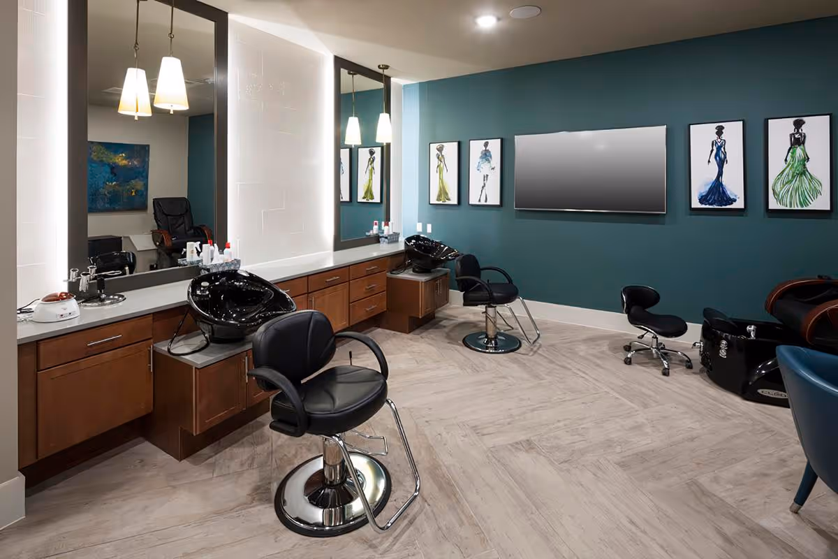 Interior view of a modern salon area with two black salon chairs in front of wash basins, large mirrors with pendant lights above, wooden cabinetry, a teal accent wall with framed fashion sketches, and a large flat-screen TV mounted on the wall.