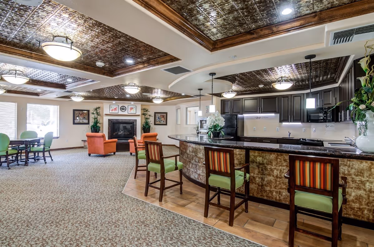 A spacious senior living facility common area featuring a kitchen with a curved counter and bar stools, a seating area with orange armchairs around a fireplace, and a dining table with green chairs near large windows. The ceiling has decorative panels and multiple light fixtures.