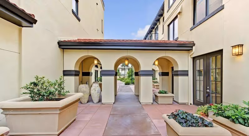 Outdoor courtyard area at Merrill Gardens at Campbell featuring a walkway with three archways, large planters with greenery, and wall-mounted lantern-style lights on a beige building under a blue sky.