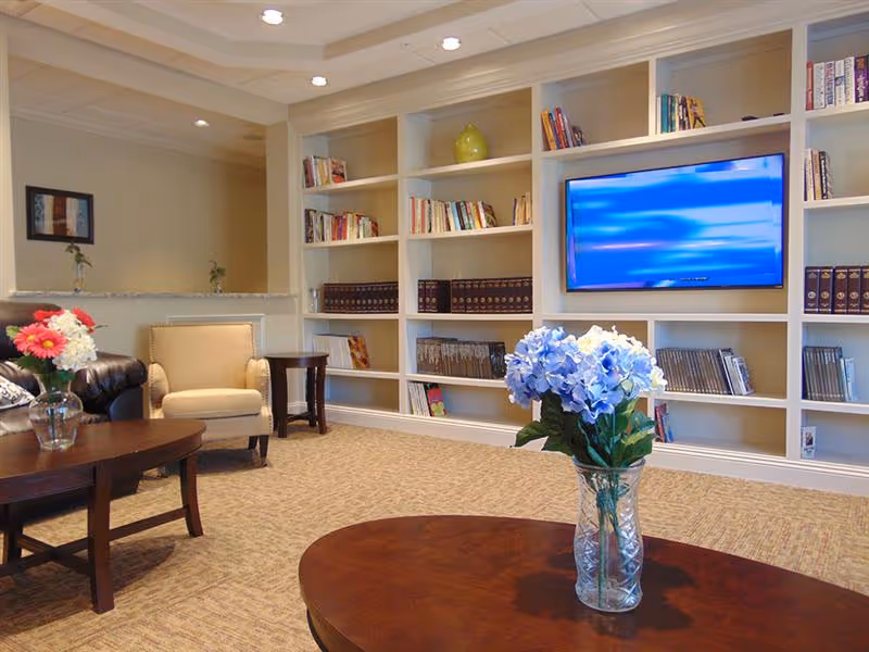 A cozy senior living common area featuring beige carpet, built-in white bookshelves filled with books and DVDs, a wall-mounted flat screen TV, a beige armchair, a dark leather sofa, and wooden coffee tables with vases of flowers.