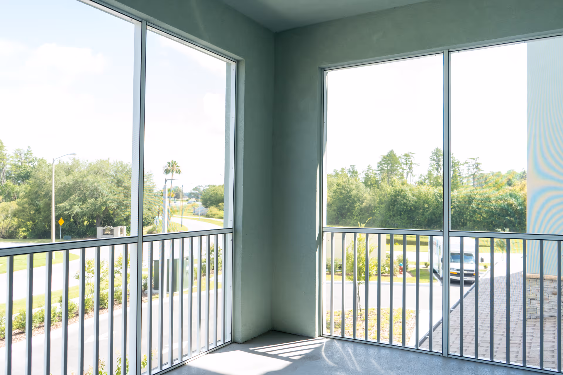 View from a screened balcony with metal railings overlooking a road, trees, and a parked white van under a bright sky.