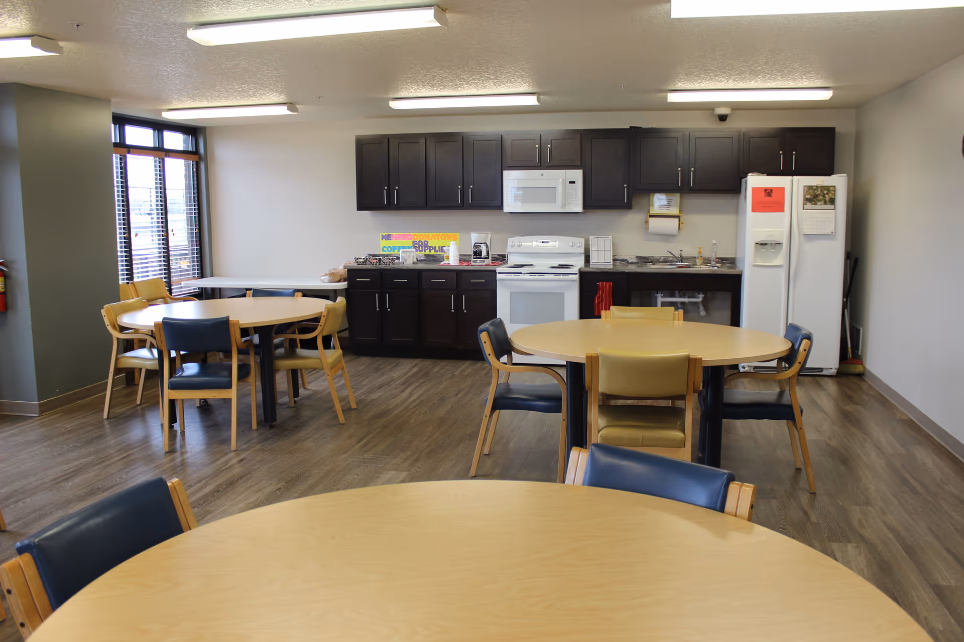 A communal kitchen and dining area with round tables and chairs. The kitchen features dark cabinets, a white stove, microwave, and refrigerator. The room has wood flooring and large windows letting in natural light.