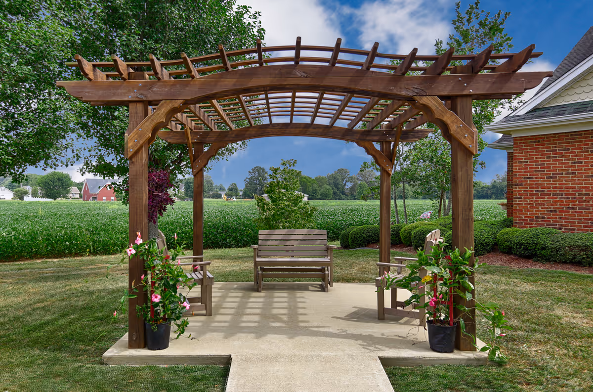 A wooden pergola with a bench and two chairs underneath it, situated on a concrete patio in a grassy outdoor area. There are potted flowering plants on either side of the pergola, green bushes, trees, and a red brick building to the right. In the background, there are open fields and a partly cloudy sky.