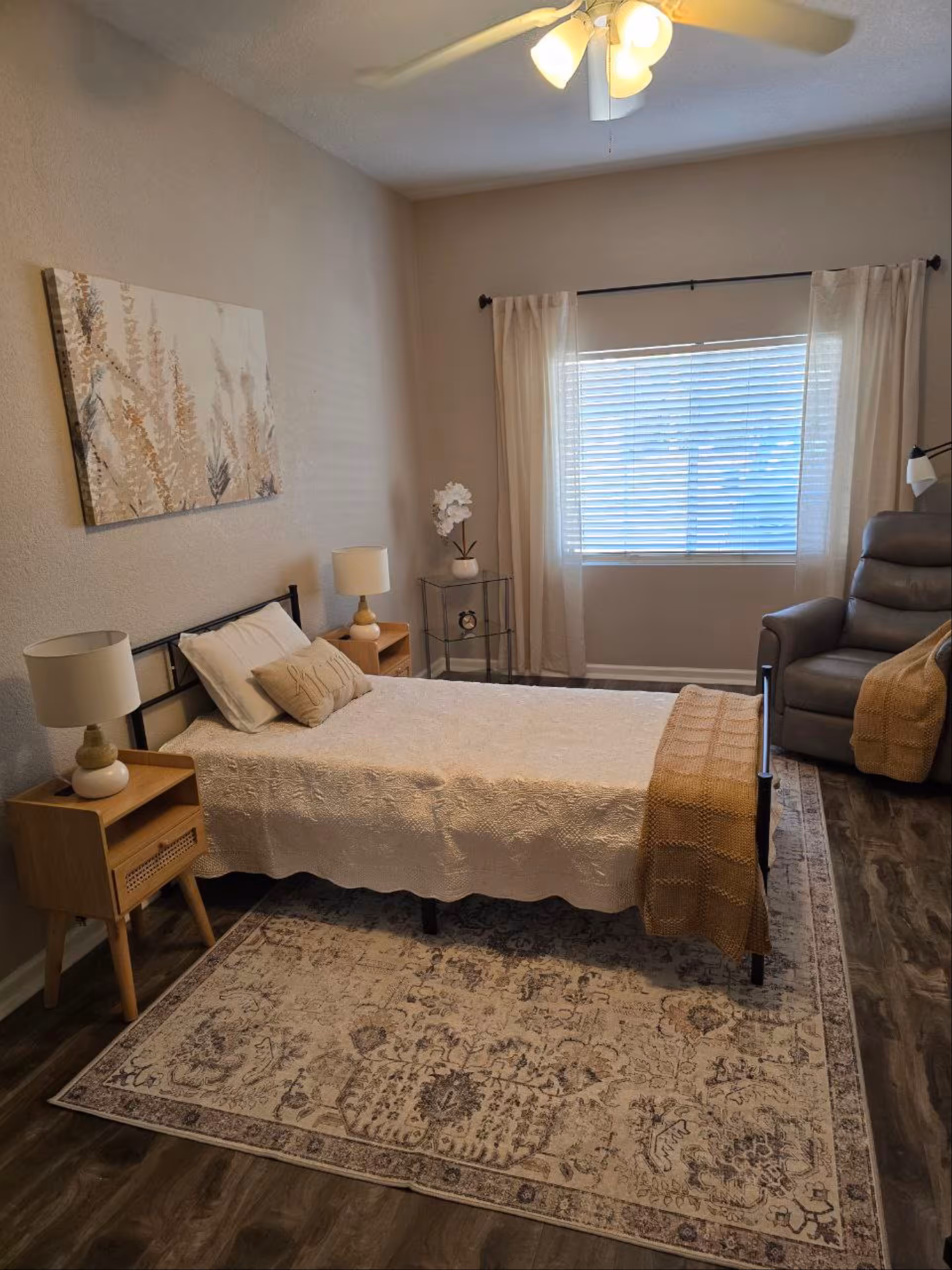 A cozy bedroom with a single bed covered in a white textured bedspread and a brown knitted throw blanket. There are two wooden nightstands with matching lamps on either side of the bed. A decorative painting hangs above the bed on the wall. A window with white curtains and blinds lets in natural light. A comfortable gray recliner chair with a brown throw blanket is placed near the window. The floor has a patterned area rug over wood-style flooring, and a ceiling fan with lights is visible above.