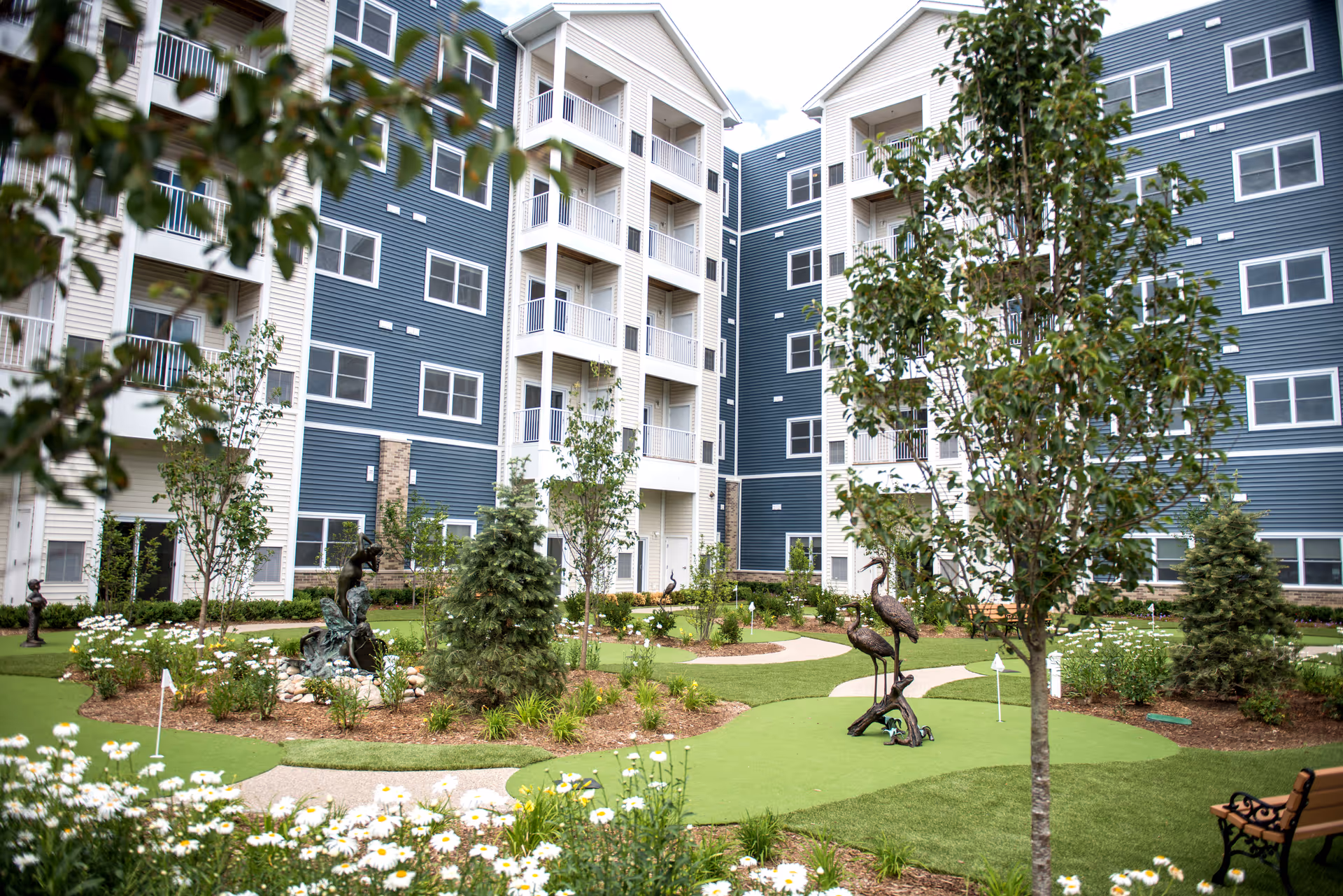 Landscaped courtyard with a small putting green, sculptures, flowers and trees in front of a multi-story blue-and-white apartment building.