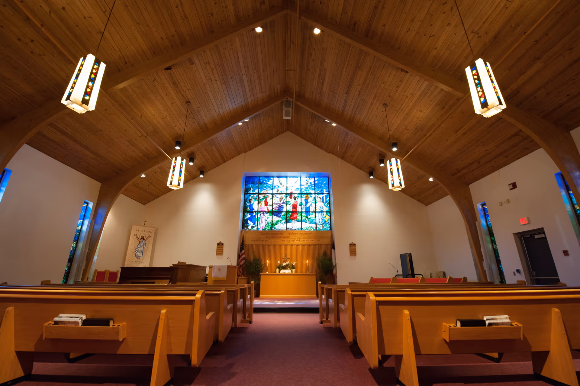 Interior view of a chapel with wooden pews, a high wooden ceiling with hanging stained glass light fixtures, and a large colorful stained glass window behind the altar.