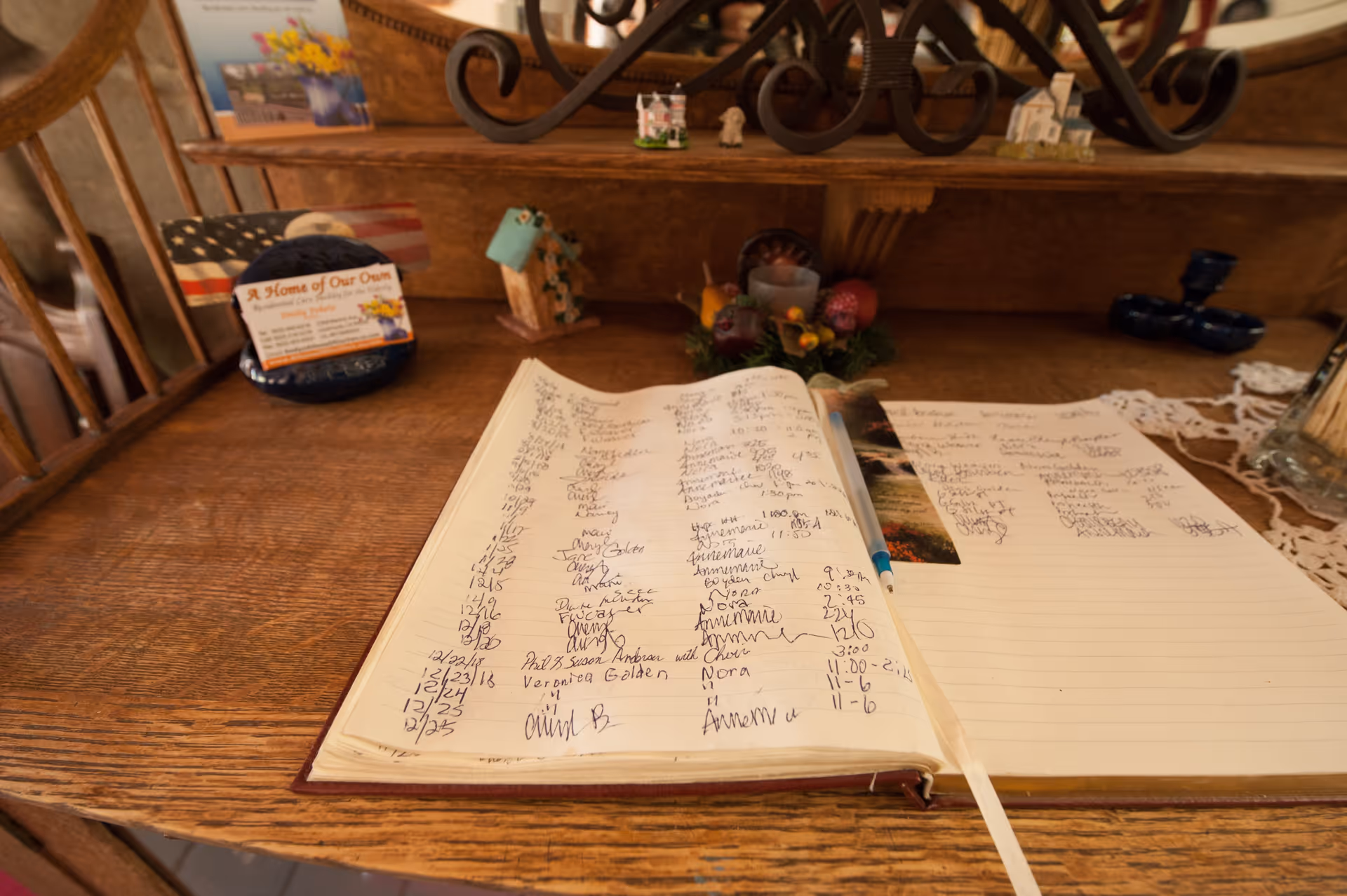 An open guestbook with handwritten notes and signatures on a wooden desk. Behind the guestbook, there are small decorative items including a miniature house, a floral arrangement, and a business card holder with a card that reads 'A Home of Our Own'.