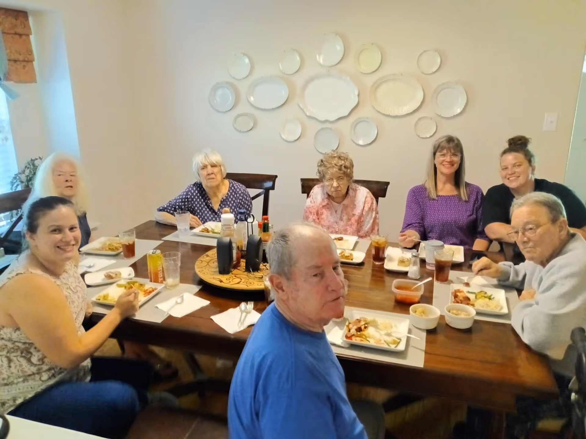 A group of seven people, including elderly individuals and caregivers, sitting around a wooden dining table enjoying a meal together in a well-lit room. The wall behind them is decorated with an arrangement of white decorative plates.