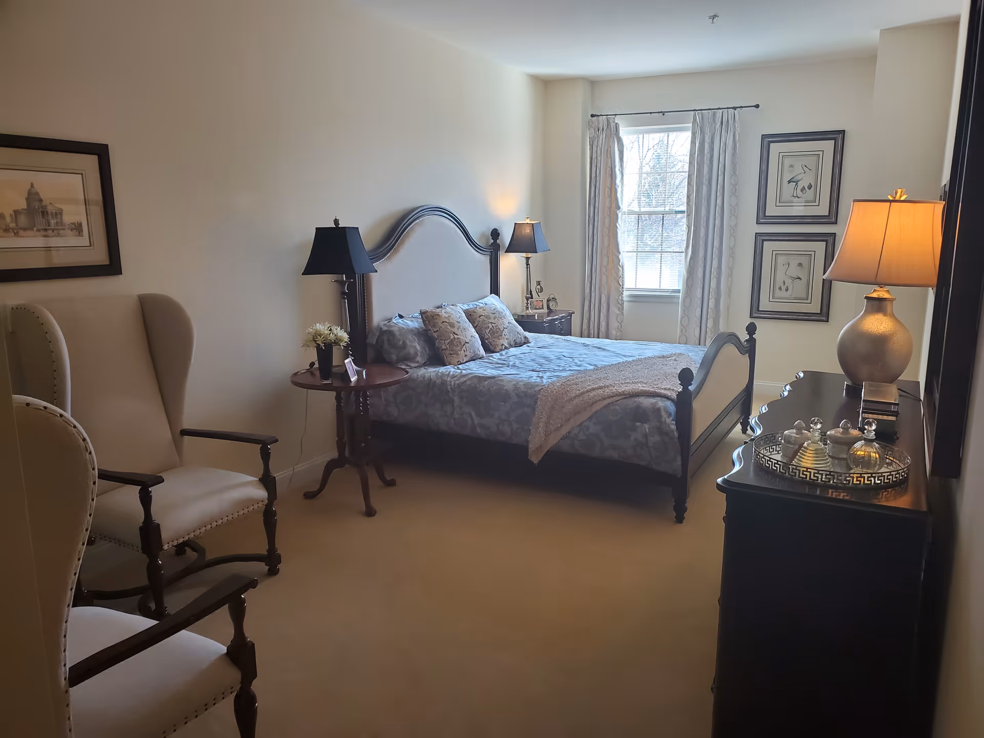 A cozy bedroom with a large bed featuring a decorative headboard and patterned bedding. Two cushioned armchairs are positioned on the left side near a small round wooden table with a lamp and a small flower arrangement. A window with light-colored curtains lets in natural light. On the right side, there is a dark wooden dresser with a lamp and decorative items on top. The walls are adorned with framed artwork.
