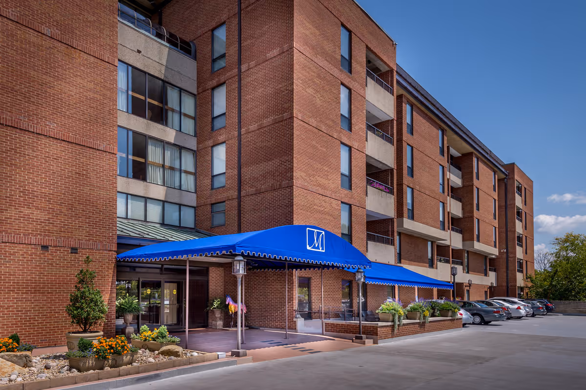 Entrance of a multi-story brick retirement community building with a blue awning, potted plants, and parked cars.