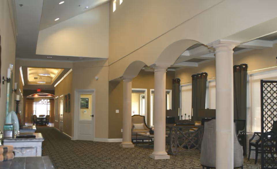 Interior view of a senior living facility hallway with beige walls, carpeted floor, and decorative columns. There are seating areas with chairs and tables along the right side, and a door and artwork on the left wall. The ceiling has recessed lighting and a high vaulted design.