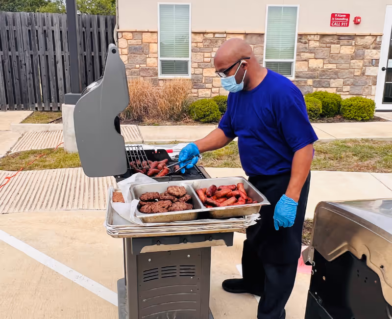 A man wearing a blue shirt, black apron, blue gloves, and a face mask is grilling hamburgers and sausages on an outdoor grill in front of a building with stone and beige walls. There are trays of cooked hamburgers and sausages on the grill's side shelf.