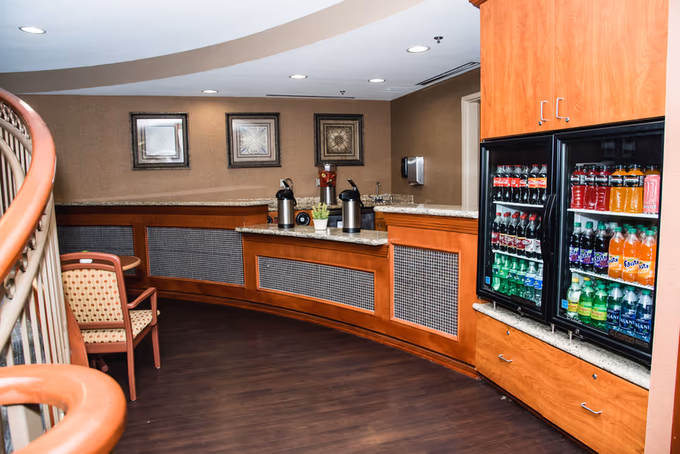 Curved common-area refreshment counter with coffee urns, a refrigerated display stocked with sodas and bottled drinks, and a nearby chair.