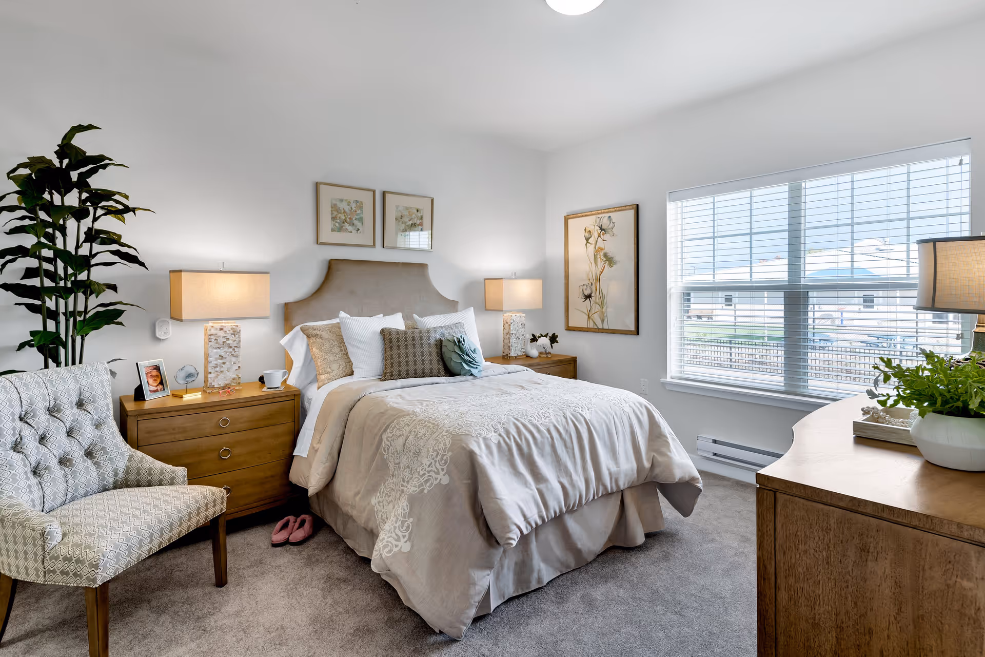 A cozy bedroom with a neatly made bed featuring beige and white bedding and multiple pillows. On either side of the bed are wooden nightstands with lamps, a photo frame, and decorative items. A patterned armchair is positioned to the left, and a large window with blinds allows natural light to fill the room. The walls are decorated with framed floral artwork, and a wooden dresser with a lamp and plant is on the right side of the room.