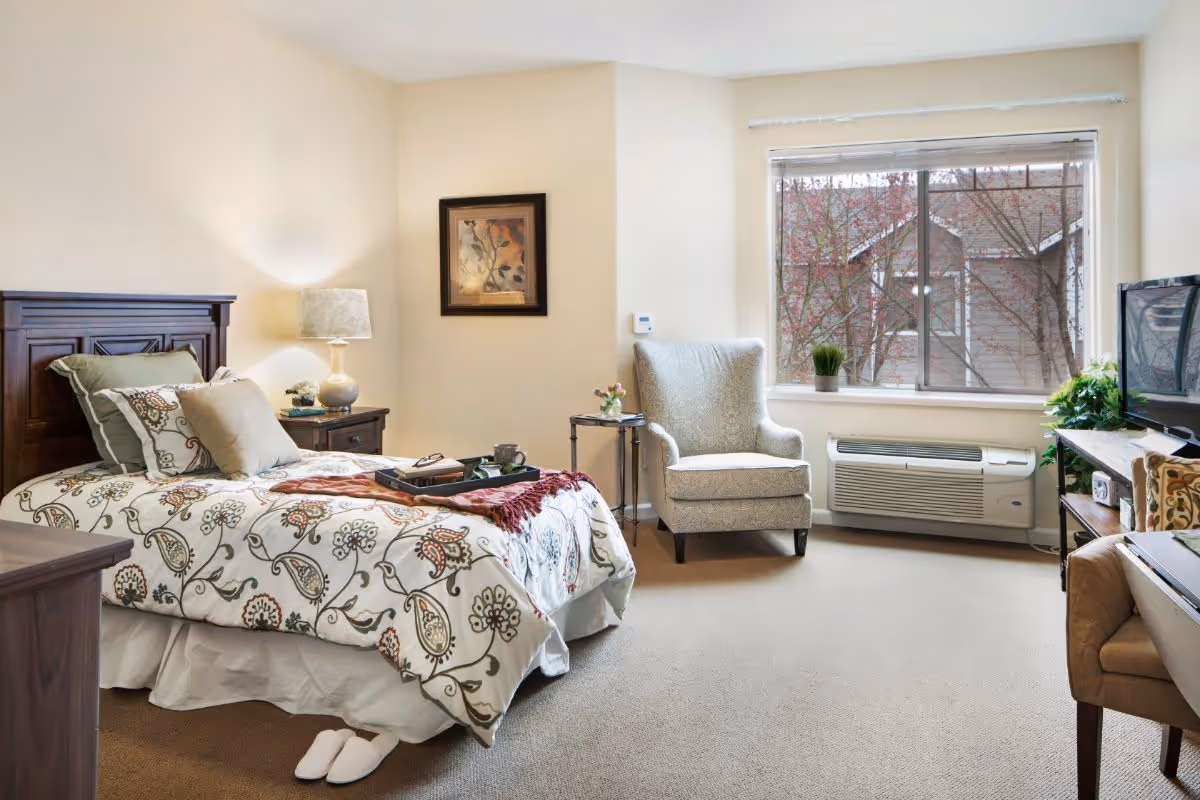 Bright, cozy bedroom with a patterned bed, nightstand and lamp, an upholstered chair by a window, and a TV.