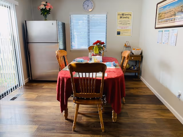 A dining area with a wooden table covered by a red tablecloth and four wooden chairs. On the table, there is a fruit basket with bananas and flowers. Behind the table is a window with blinds, a clock above it, and a refrigerator to the left. The room has wooden flooring, a small side table with items on it, and a framed picture on the wall.