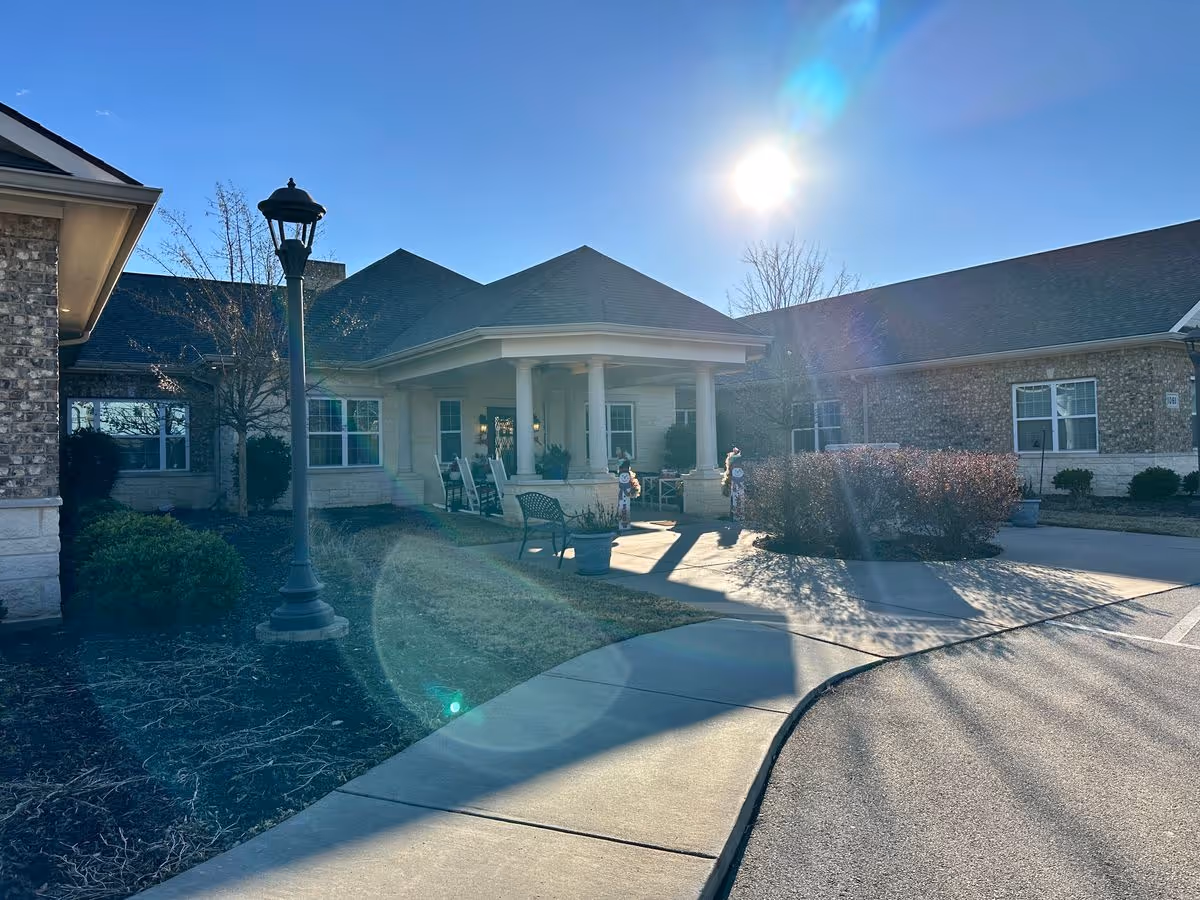 Covered front entrance of a single-story brick senior living building with outdoor seating, a lamp post, and a curved driveway under bright sun.
