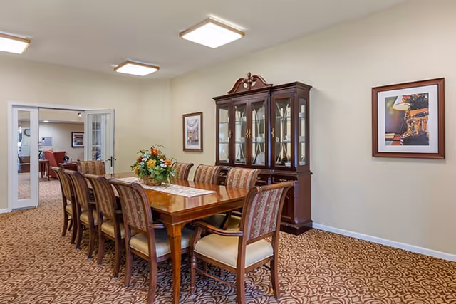 A dining room with a long wooden table surrounded by eight upholstered chairs. A floral centerpiece is on the table. Behind the table is a dark wooden china cabinet with glass doors displaying decorative items. The walls are light-colored and decorated with framed artwork. The floor is carpeted with a patterned design. In the background, there is an open doorway leading to another room with additional seating.