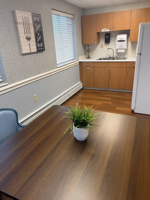 A small kitchen area with wooden cabinets, a sink, a white refrigerator, and a paper towel dispenser mounted on the wall. In the foreground, there is a wooden table with a small potted plant on it and a blue chair next to the table. The walls are light gray with a framed artwork hanging near a window with white blinds.