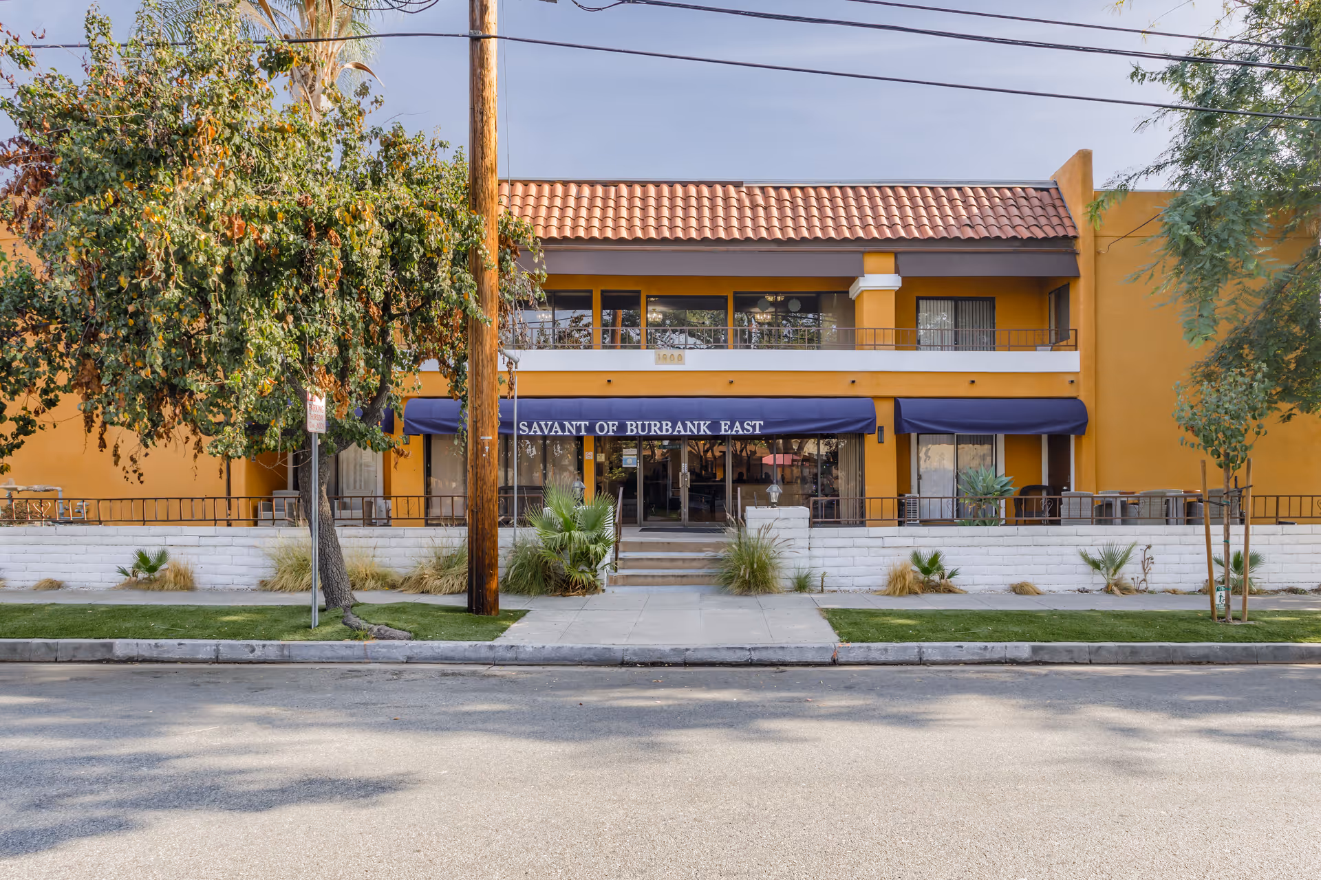 Front exterior view of Savant of Burbank East, a two-story building with a tiled roof and yellow-orange walls. The entrance has a blue awning with the facility name, and there are trees and landscaping in front along the sidewalk.
