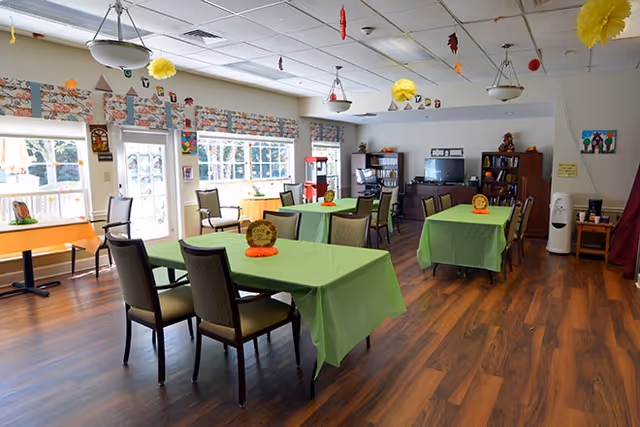A bright and spacious dining room with wooden floors and several tables covered with green tablecloths. Each table has chairs around it and small decorative centerpieces. The room has large windows letting in natural light, ceiling lights, and colorful hanging decorations. In the background, there is a TV, bookshelves, and a popcorn machine near the windows.