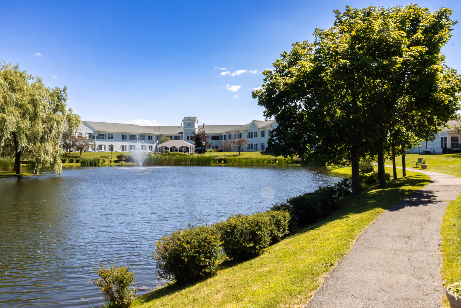 A large pond with a fountain in the center, surrounded by green grass, bushes, and trees. A paved walking path runs along the right side of the pond. In the background, there is a two-story white building with many windows under a clear blue sky.