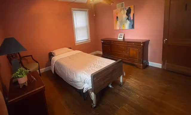 Single-occupancy bedroom with a twin bed on wheels, wooden dresser, side tables, a chair, ceiling fan, and pink walls.