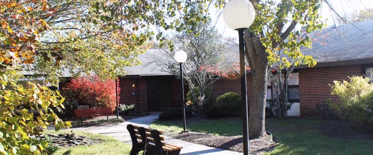 Outdoor view of a senior living facility courtyard with a paved walkway, benches, lamp posts, trees with autumn foliage, and a brick building in the background.
