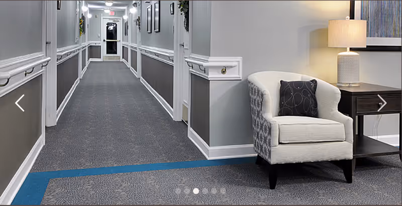 A long, well-lit hallway in a senior living facility with gray walls and patterned carpet. On the right side near the foreground, there is a beige upholstered armchair with a dark decorative pillow next to a wooden side table with a lamp and framed artwork above it.