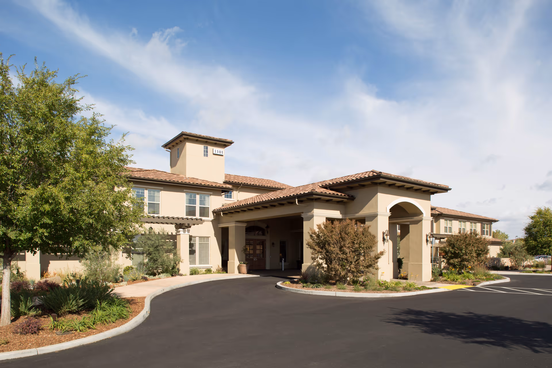 Exterior view of a senior living facility building with beige walls and a tiled roof, surrounded by landscaped greenery and trees under a partly cloudy sky.
